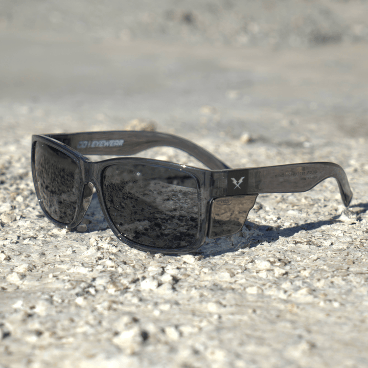 CO Eyewear Oiler Z87+ Safety Glasses with Gray Frame and Smoke Lenses laying on gravel